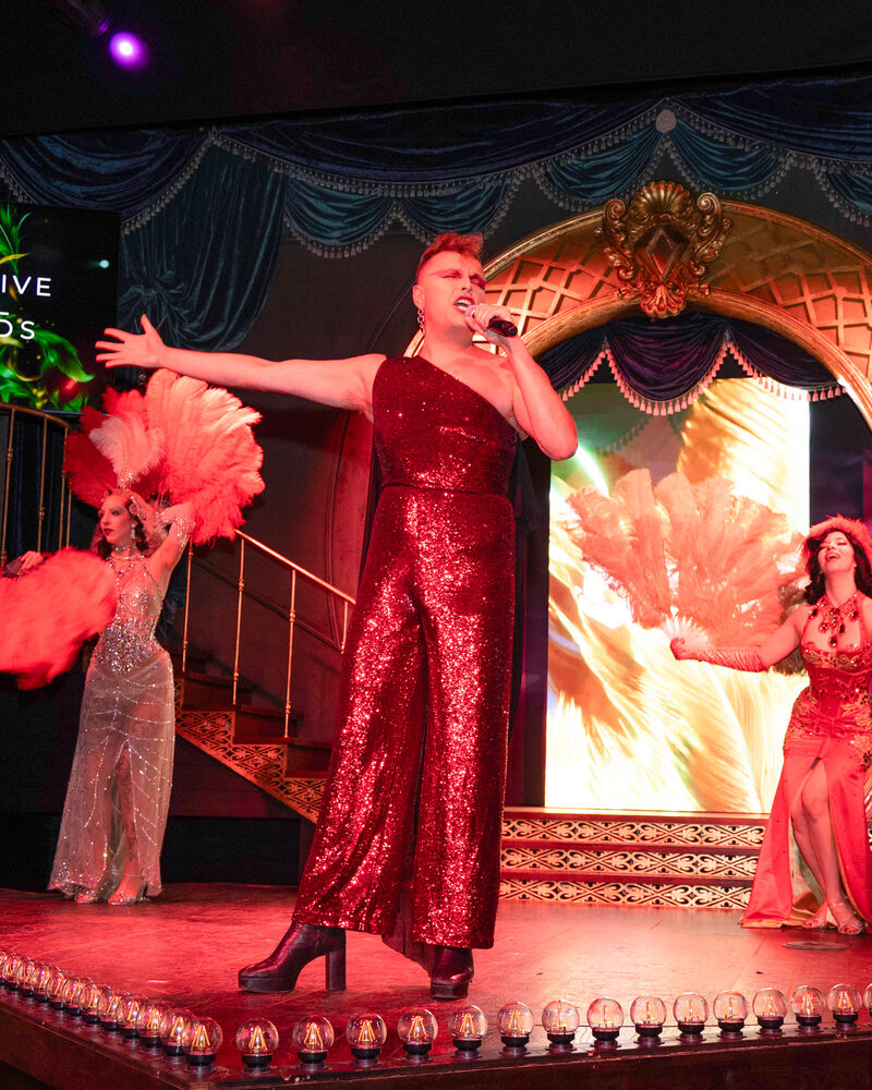 A performer in red sequins singing on stage at the Emerald Theatre, with two fan dancers